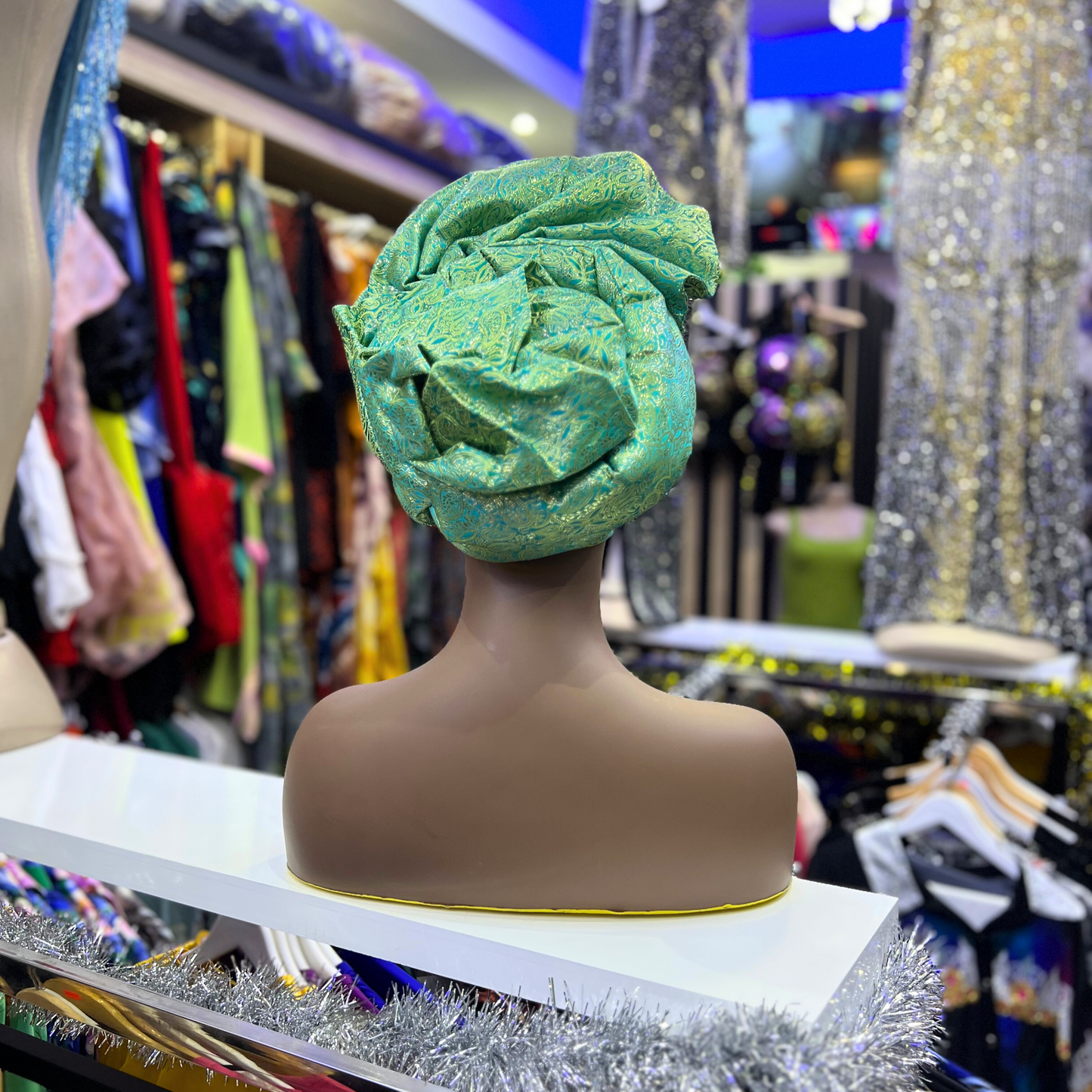 Draped Gele head wrap in vibrant green, styled elegantly on a mannequin with colorful clothing in the background